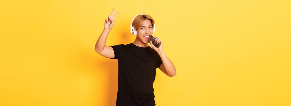 Sassy handsome asian guy in headphones, playing karaoke app, singing into mobile phone microphone, showing peace gesture, standing yellow background.