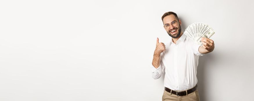 Successful businessman showing money dollars and thumbs-up, smiling satisfied, standing over white background. Copy space