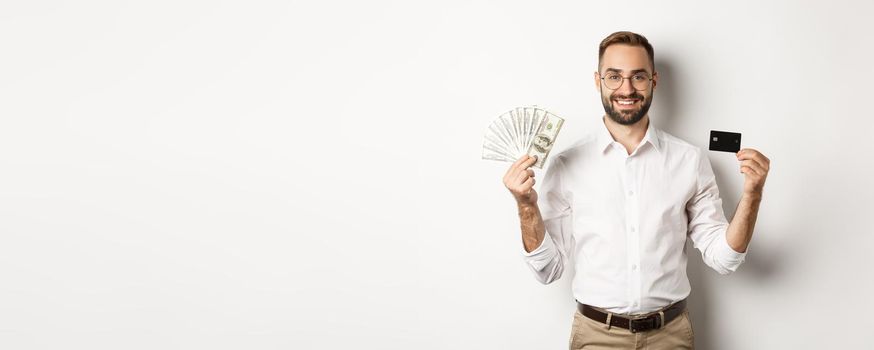 Handsome business man showing credit card and money dollars, smiling satisfied, standing over white background.