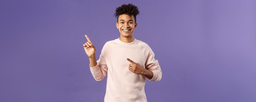 Portrait of cheerful, healthy smiling man pointing fingers upper left corner, recommending buy product, shop online, order delivery, advertisement and people concept, purple background.