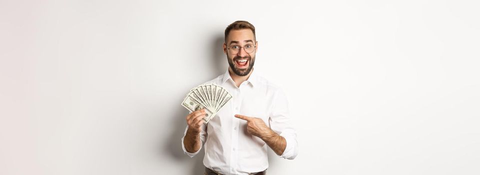 Excited businessman pointing at money, showing dollars and smiling, standing over white background.