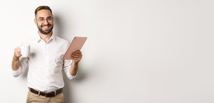 Satisfied boss drinking tea and using digital tablet, reading or working, standing over white background.