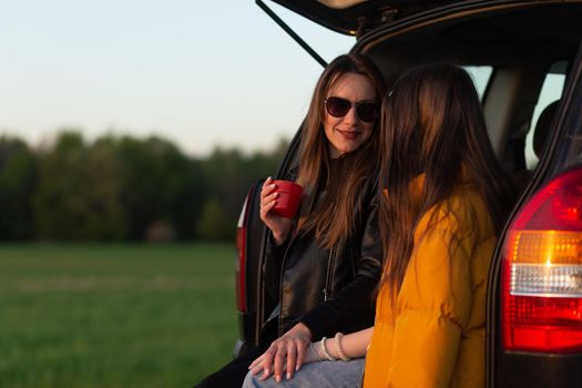 Mother and daughter camping on a hill and admiring the sunset while sitting in the car trunk