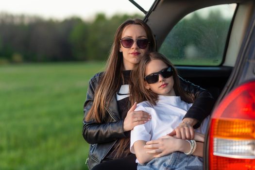 Mother and daughter camping on a hill and admiring the sunset while sitting in the car trunk