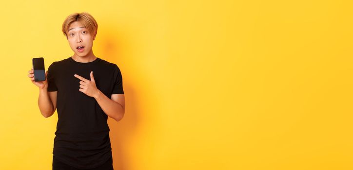 Portrait of handsome asian man showing something on smartphone screen, pointing at mobile phone display, standing over yellow background.