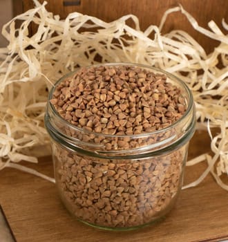 brown raw buckwheat in a glass pot stands on a wooden table against a background of paper shavings. vegetarian food. healthy carbohydrates. High quality photo