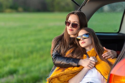 Mother and daughter camping on a hill and admiring the sunset while sitting in the car trunk