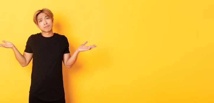 Portrait of clueless asian blond guy, wearing black clothes, shrugging and looking puzzled, standing yellow background.