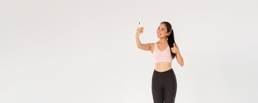 Full length of smiling female lifestyle blogger, asian fitness girl in sportswear taking selfie in gym, showing thumbs-up, demonstrate workout exercies for followers online, live stream from training.