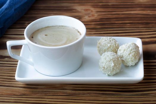 a cup of frothy latte stands on a square saucer on a wooden table with coconut sweets. Coffee with dessert. High quality photo