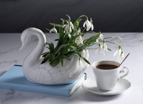 Still life with white snowdrops in a ceramic vase in the form of a swan, coffee in a white cup, conceptual morning ritual whith a book.High quality photo