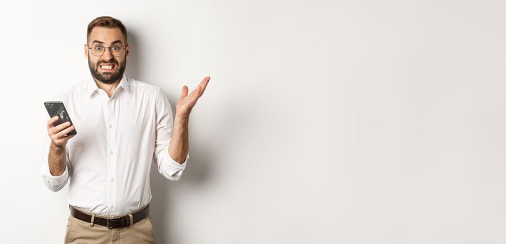 Confused businessman reading strange message on mobile phone, looking annoyed, standing over white background.