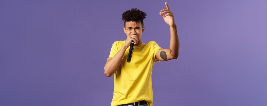 Portrait of young carefree sassy hipster guy with tattoos, yellow t-shirt performing in front audience with his hip-hop or rap, singing song, raising finger up, hold microphone, karaoke.