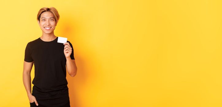 Cheerful handsome asian guy smiling pleased and showing credit card, standing yellow background.