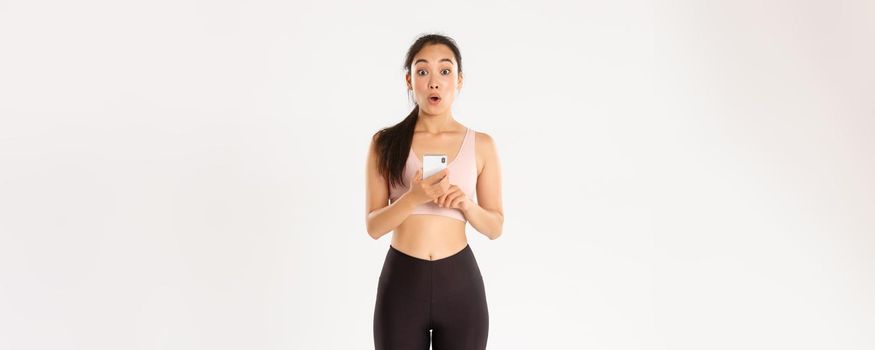 Sport, technology and active lifestyle concept. Surprised and amazed asian female athlete, sportswoman say wow, checking fitness app on mobile phone, using workout tracker, running with application.