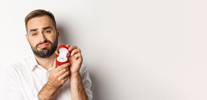 Close-up of hopeful man begging to marry him, looking sad and showing wedding ring, making a proposal, white background.