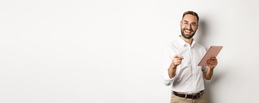 Happy and satisfied boss praising good job, reading on digital tablet and pointing at you camera, standing over white background.