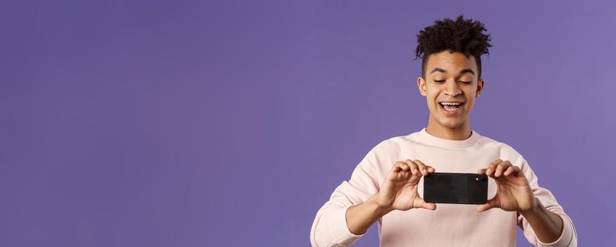 Portrait of amazed, excited young man seeing something interesting, stream concert to his internet social network profile, taking photo or recording video with mobile phone, purple background.