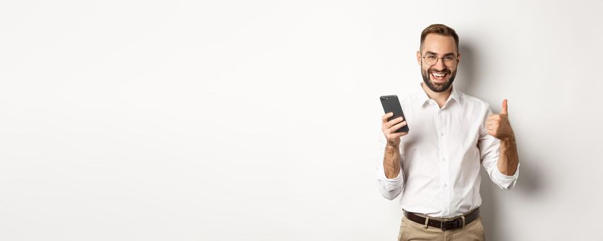 Satisfied business man showing thumbs up after using mobile phone, standing pleased over white background.