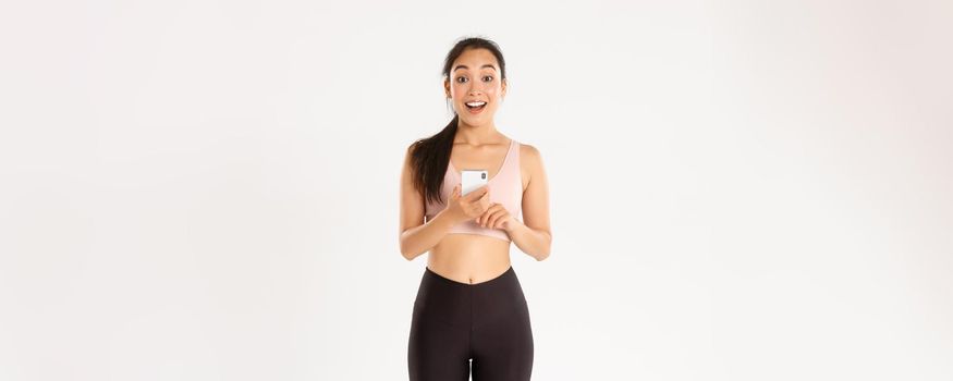 Sport, technology and active lifestyle concept. Surprised and amazed asian female athlete, sportswoman say wow, checking fitness app on mobile phone, using workout tracker, running with application.