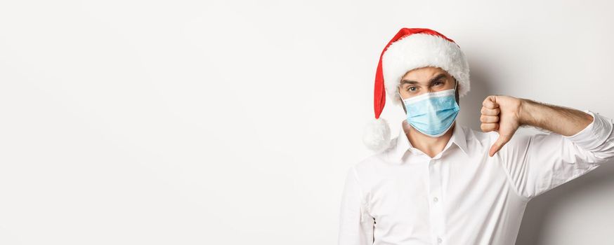 Concept of covid-19, social distancing and winter holidays. Close-up of disappointed man in face mask and christmas hat showing thumb down, standing over white background.