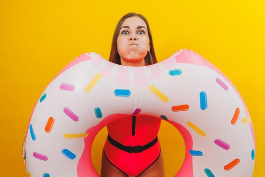 Emotional young woman wearing a pink swimsuit with an inflatable ring isolated on a bright yellow background. Summer hotel pool sea vacation sun tanning concept