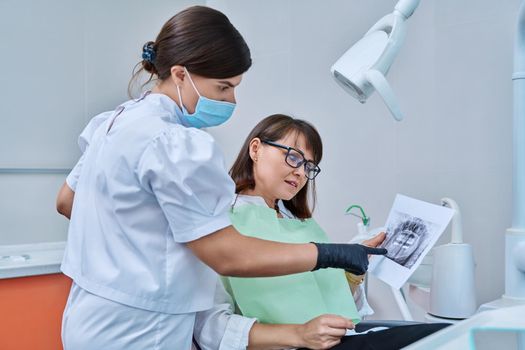 Female doctor dentist talking to middle aged woman patient in dental chair, discussing x-rays of teeth and jaws. Dental treatment and prosthetics, implantation, health care concept