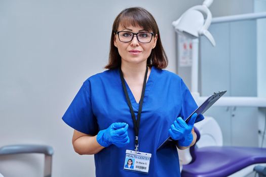 Portrait of female dentist in office. Middle aged doctor nurse looking at camera with professional badge clipboard near dental chair. Dentistry, medicine, specialist, career, dental health care concept