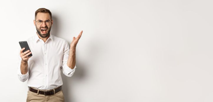 Frusteated man using mobile phone and looking disappointed, complaining, standing over white background.