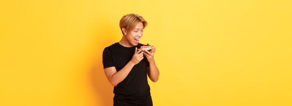 Joyful handsome asian guy smiling and looking tempted at cake while making bite, standing yellow background.