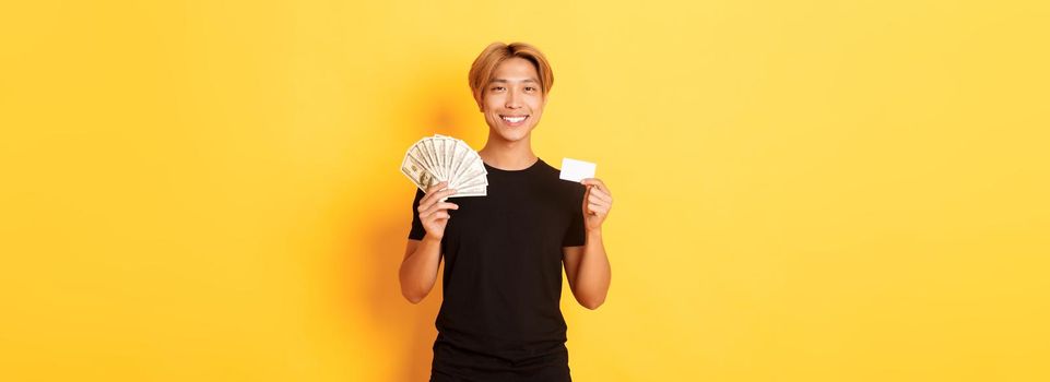 Pleased handsome asian guy showing money and credit card, smiling happy, standing yellow background.