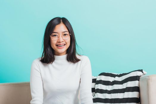 Asian beautiful young woman wearing eyeglasses sitting on sofa looking at camera, portrait relaxation of happy female smiling in living studio shot isolated on blue background