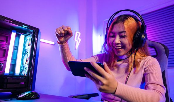 Winning. Happy Gamer playing video game online with smartphone with neon lights she raises hands to wins celebrating, confused Asian woman wearing gaming headphones feeling excited at home