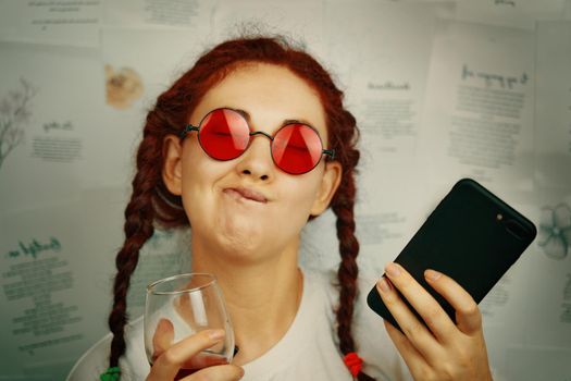 Female blogger with a glass of wine and a smartphone in her hand. Funny girl with pigtails and red sunglasses makes faces. Pampering and antics. Hippie or boho style fashion blog.