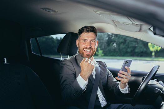 Male businessman successful rejoices and smiles reads news from mobile phone win and success meeting, man sits in car
