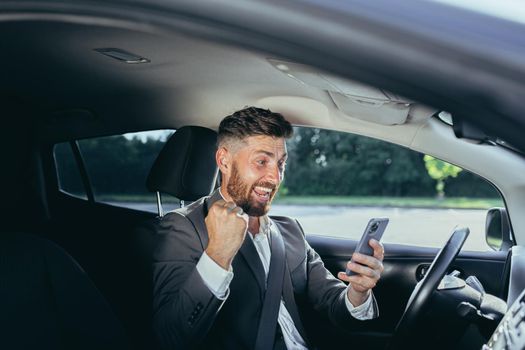 Excited emotional adult businessman driving car rejoicing online victory achievement lottery celebrating success screaming funny enjoying. Man holding his smartphone feeling happy Transportation.