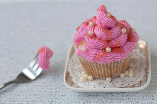 a cupcake beautifully decorated with pink cream and sugar decor stands on a square saucer on a light wooden background. A piece of muffin on a fork nearby. Sugar beads and fruit topping