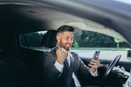 Male businessman successful rejoices and smiles reads news from mobile phone win and success meeting, man sits in car