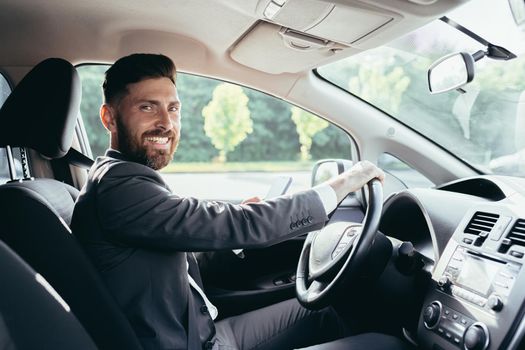 Young man with a beard businessman driving a car in the parking lot, looking at the camera and smiling