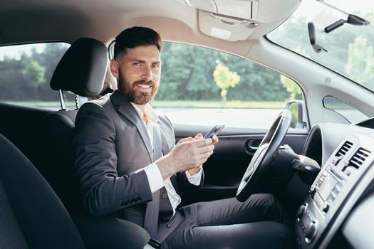 Young man with a beard businessman driving a car in the parking lot smiles and writes on the phone reads the news successful man