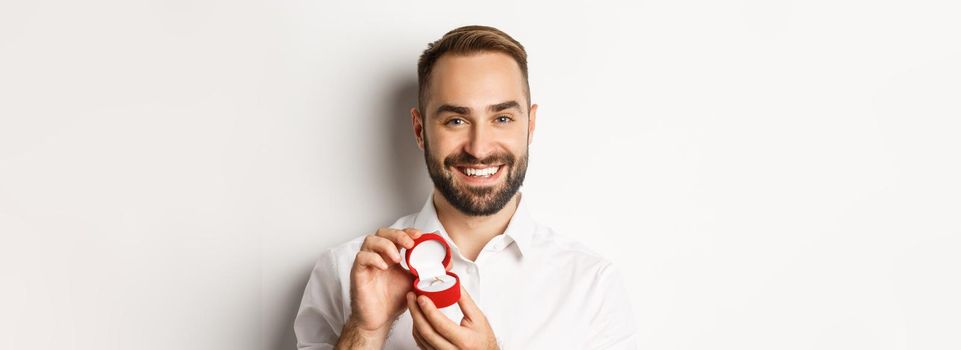 Close-up of happy handsome man making a proposal, holding wedding ring in box and smiling, asking to marry him, white background.