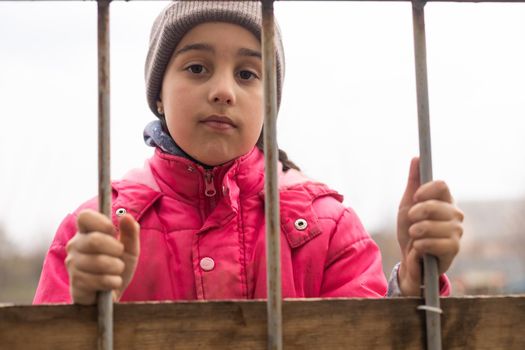 A little girl with a sad look behind a metal fence. Social problem of refugees and forced migrants.