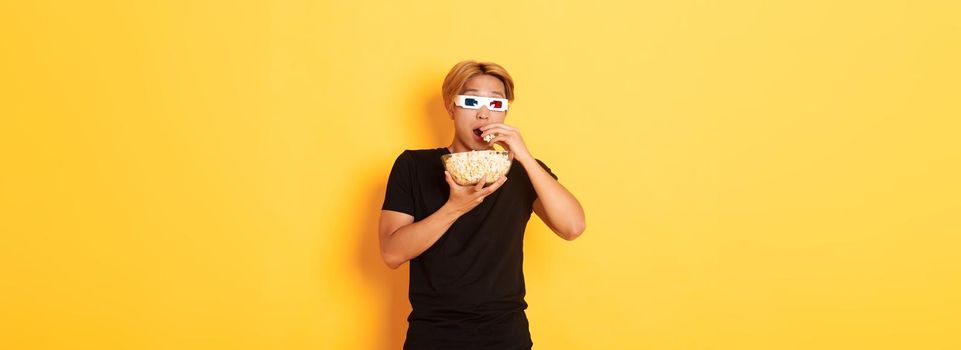 Impressed and thrilled asian guy attend cinema, watching movie or tv series in 3d glasses, eating popcorn and looking shocked, standing yellow background.