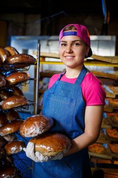 Smiling baker girl in apron holding bread and looking at camera. Young woman is a chef holding fresh baking. A happy girl of European appearance is smiling at the bakery.