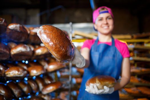 Smiling baker girl in apron holding bread and looking at camera. Young woman is a chef holding fresh baking. A happy girl of European appearance is smiling at the bakery.