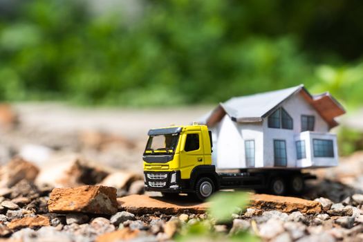 Close-up of a small toy truck on a tree stump carrying a miniature house model on top against a blurred forest background, front view. Countryside relocation service concept