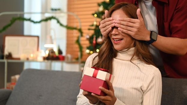 Surprised young woman sitting on the couch and holding a gift box while her boyfriend standing behind and covering her eyes with hands.