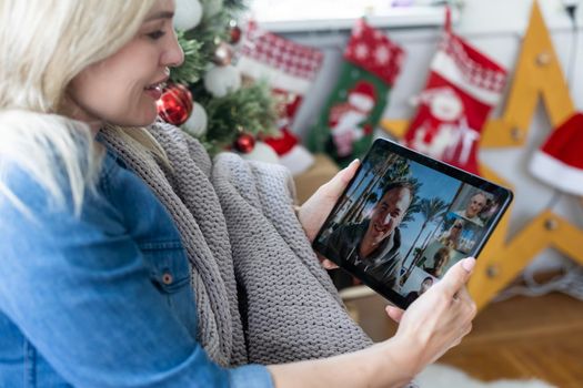 A young woman on the background of a Christmas tree with gifts, with a tablet has a video call or video chat with friends, family. Christmas online holiday. Stay home vacation.