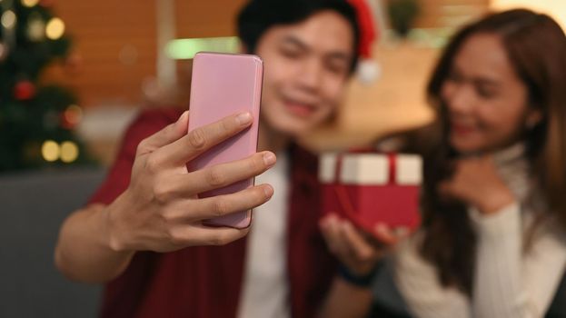 Happy young couple in Santa hat taking selfie on mobile phone while sitting in decorated room. Celebration, holidays and people concept.