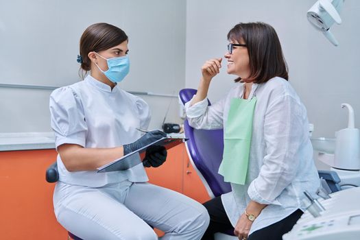 Dentist and woman patient in dental clinic, doctor nurse writing on clipboard consulting, middle aged female talking about disturbing dental problem. Dentistry, treatment, dental health care concept